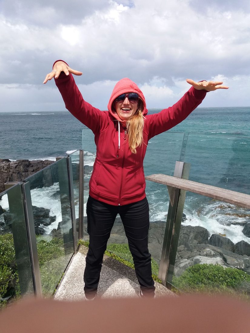 Nathalie Sameli im roten Pullover mit ausgebreiteten Armen auf einer Aussichtsplattform am Meer